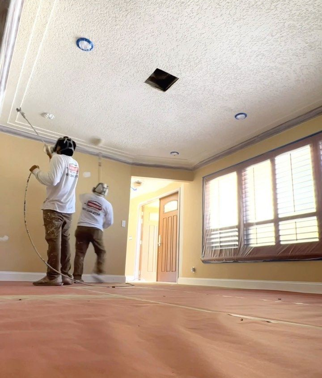 Two workers painting a room, with a ladder, window blinds, and a ceiling repair opening.