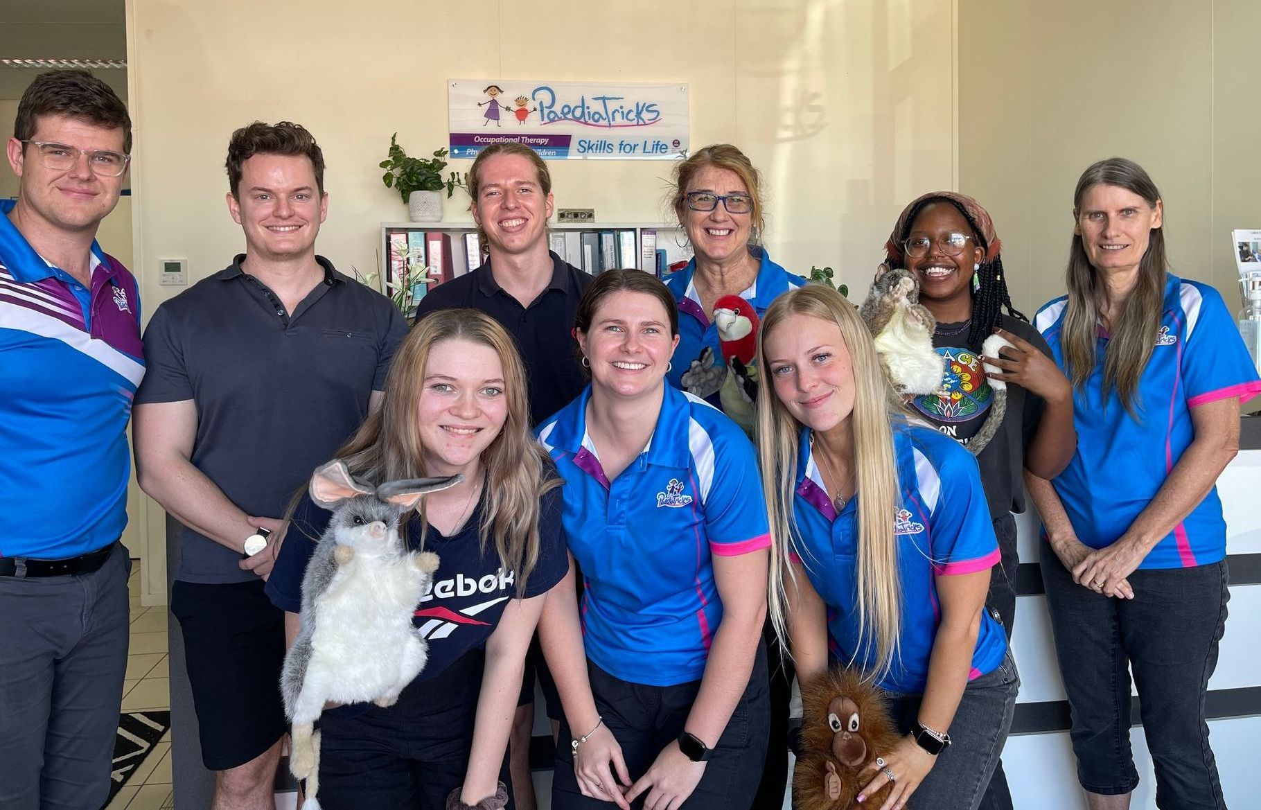 Group of people in paediatricks shirts posing indoors with puppets in front of a banner