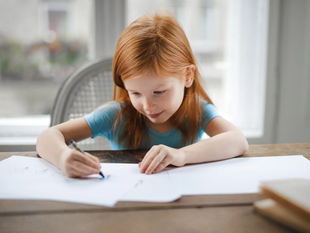 Child with red hair drawing on paper at a table, focused in a bright room