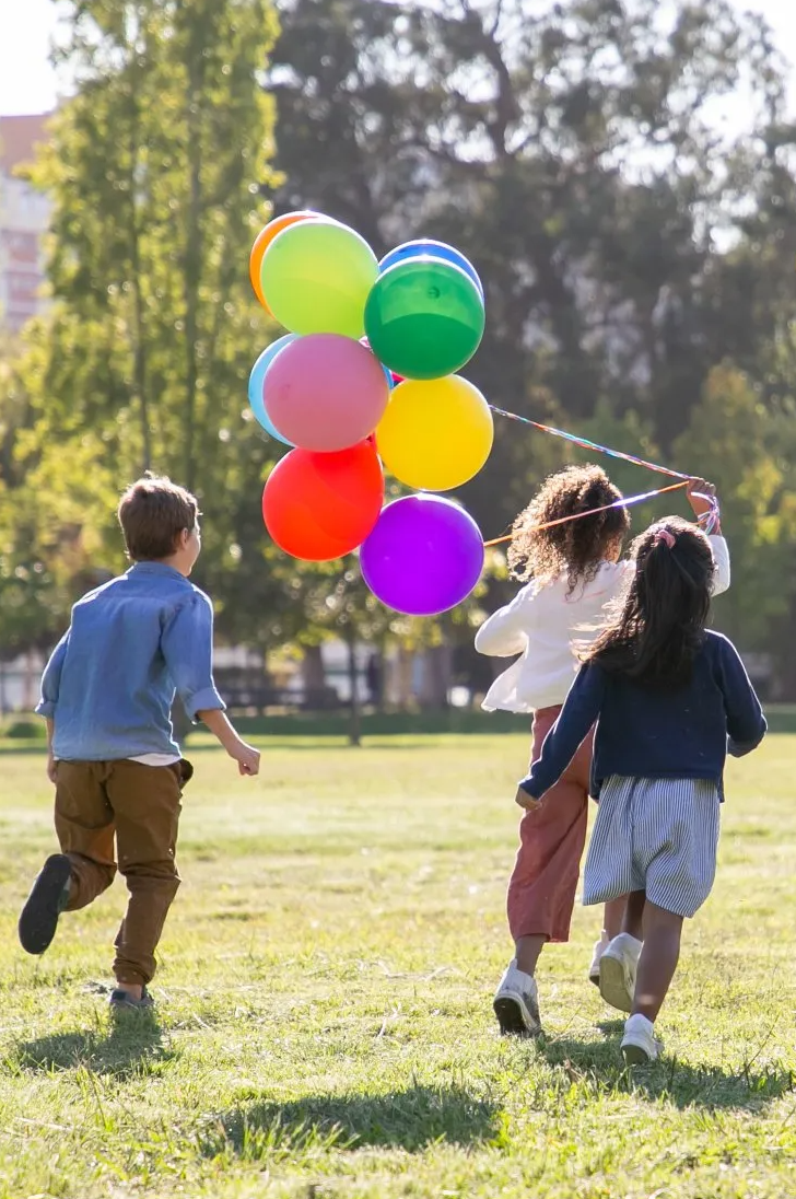 Children running toward a cluster of colorful balloons in a sunny park