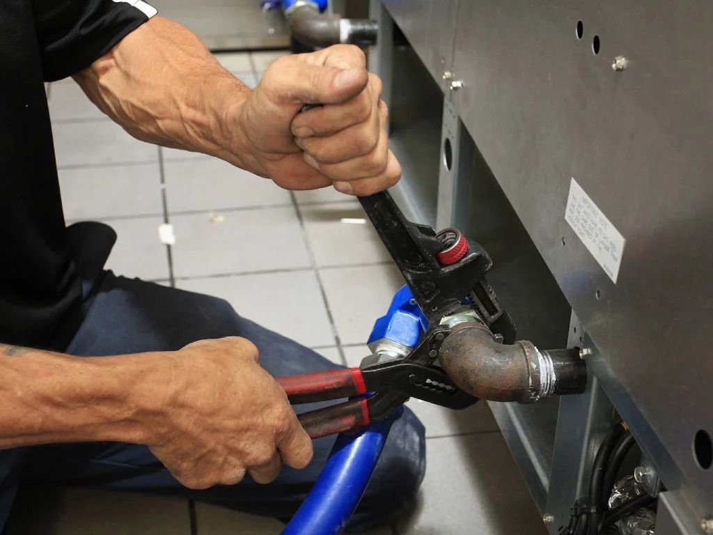 Hands using pliers to tighten a pipe fitting on metal machinery.