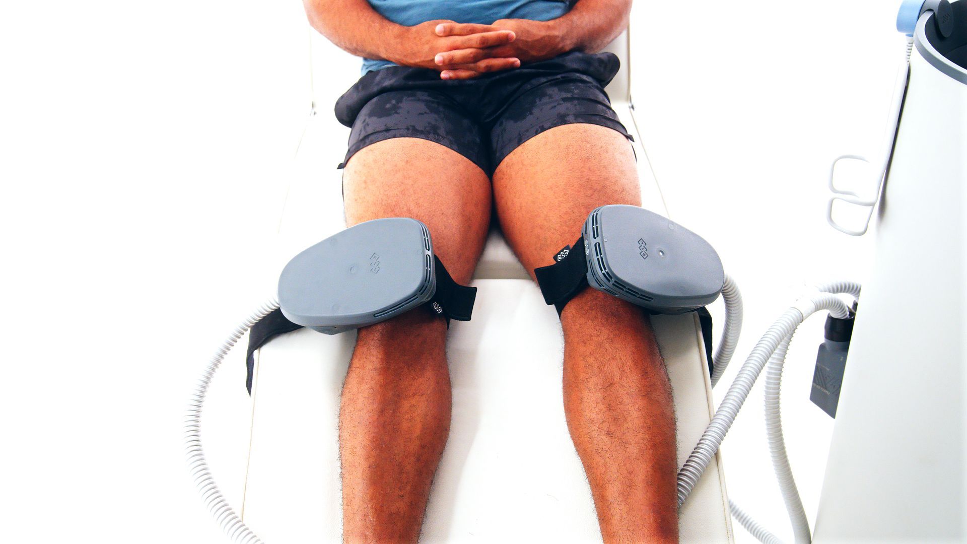 Person seated with gray knee braces attached by hoses, likely in a medical treatment setting