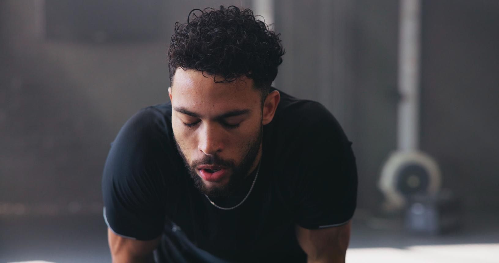 Man in a black shirt leaning forward in a dim gym, looking down and breathing heavily