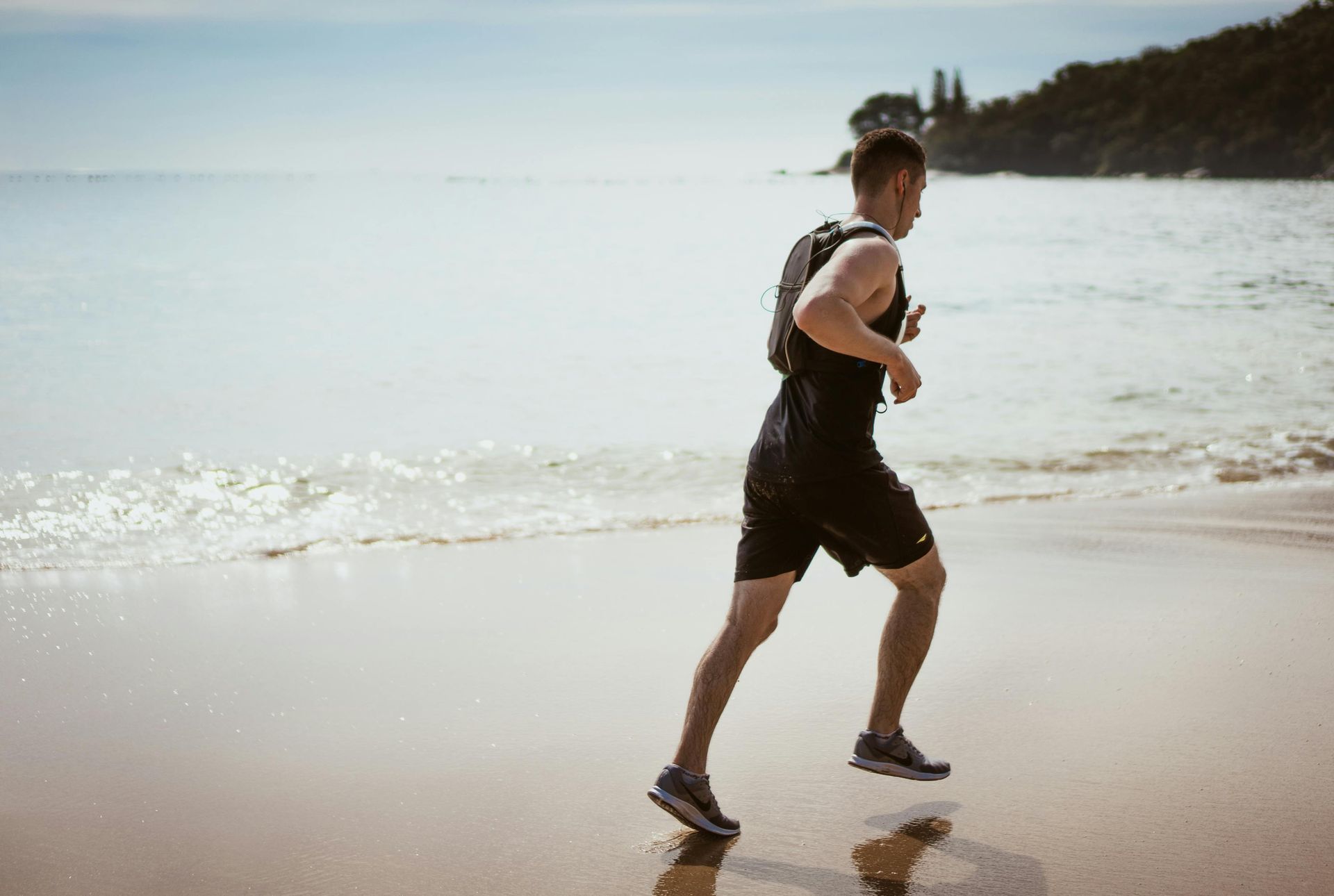 Person running on a sandy beach near the ocean, wearing black shorts and a backpack.