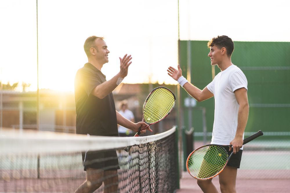 Two tennis players high-fiving over the net after a match, backlit by the sun.
