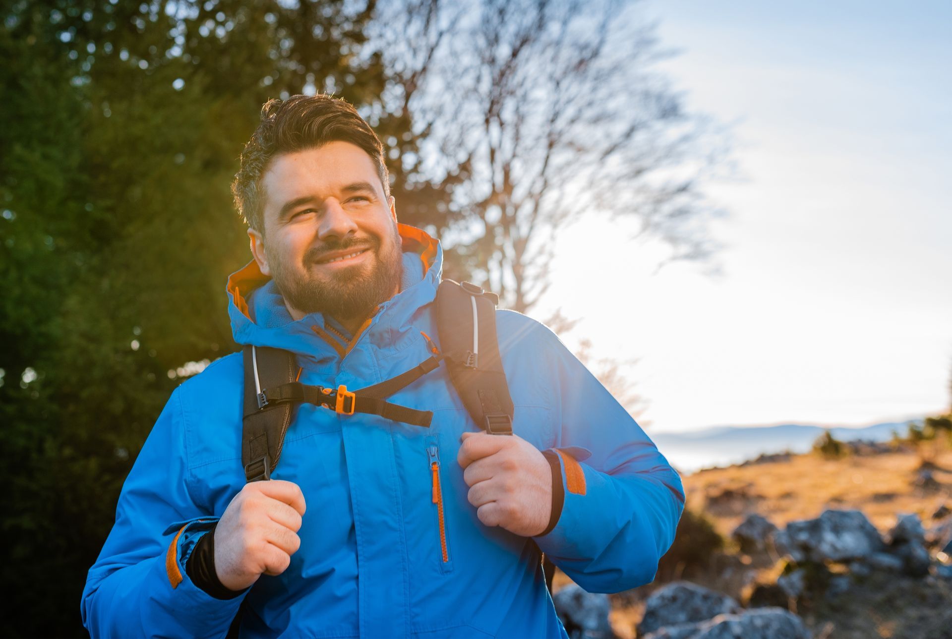 Man with backpack smiles, hiking outdoors in blue jacket, sunny day.