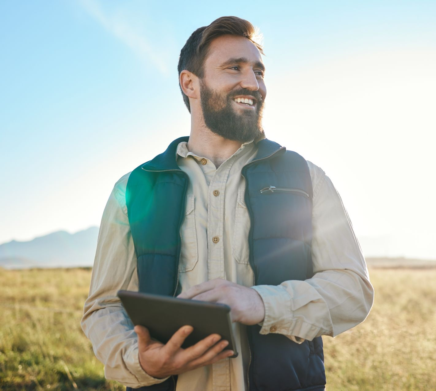 Man in field holding tablet, smiling, wearing vest.