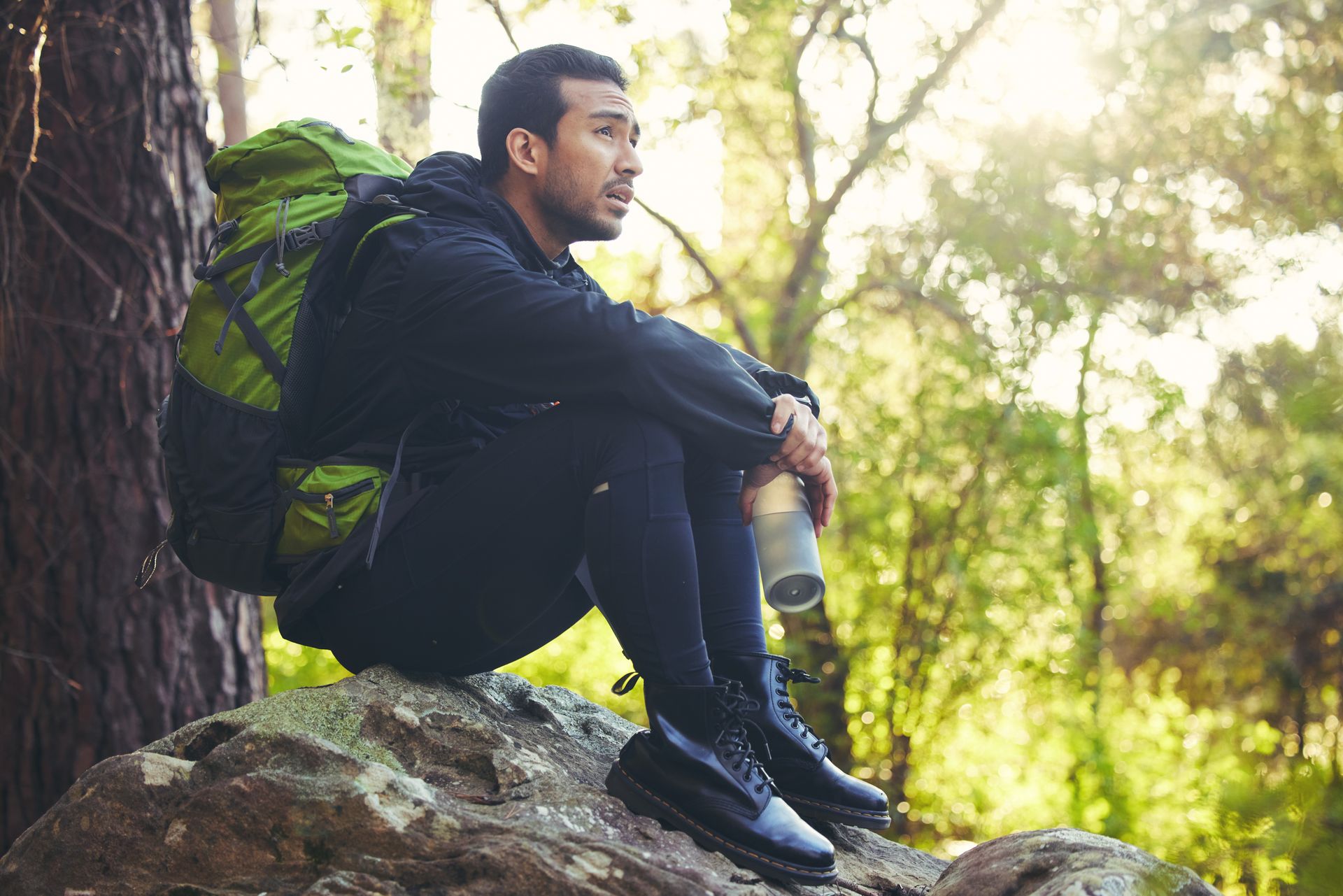 Man with backpack sitting on a rock in a forest, holding a water bottle, gazing into the distance.
