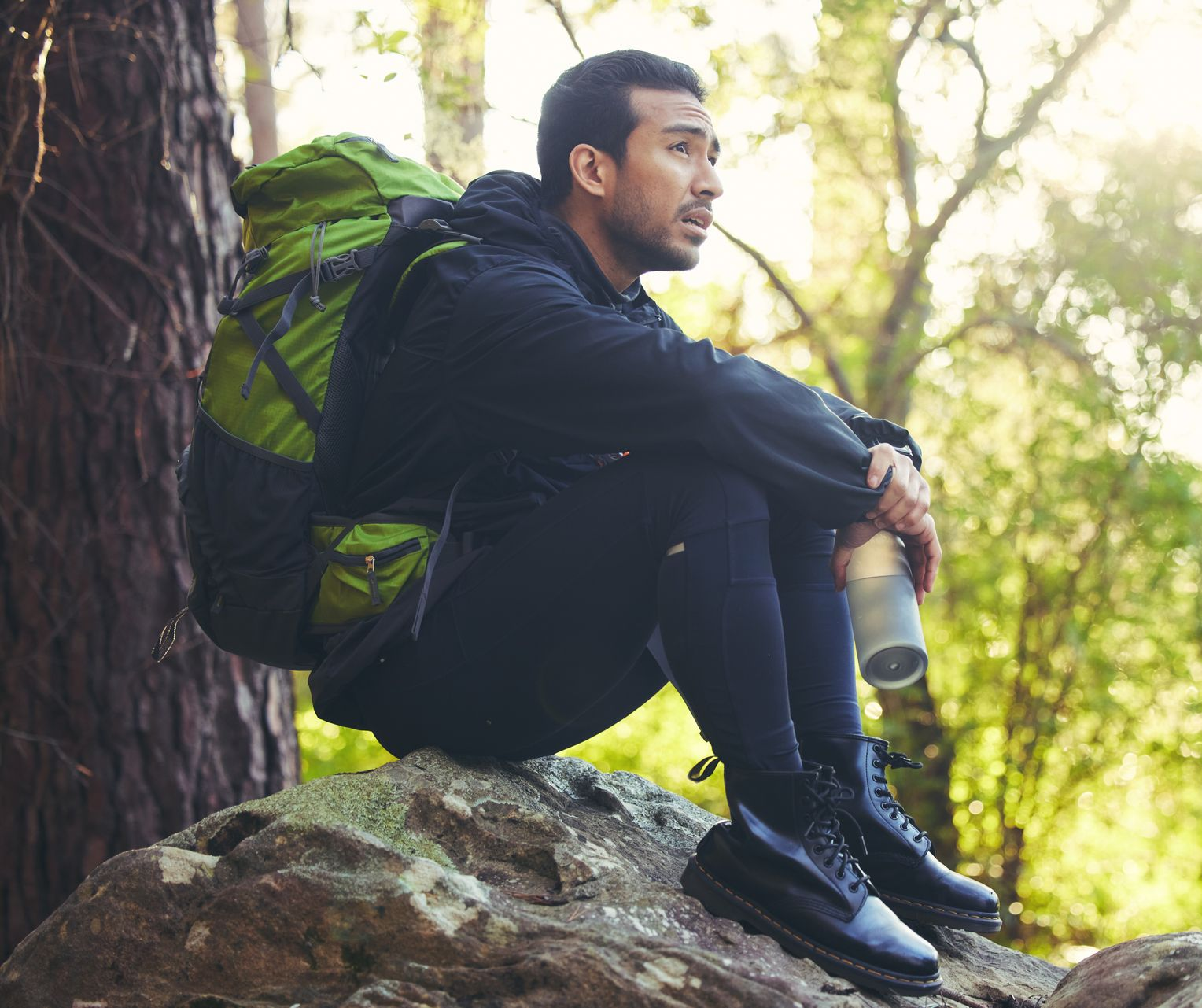 Man with backpack, resting on a rock in a forest, holding a water bottle, looking off to the side.
