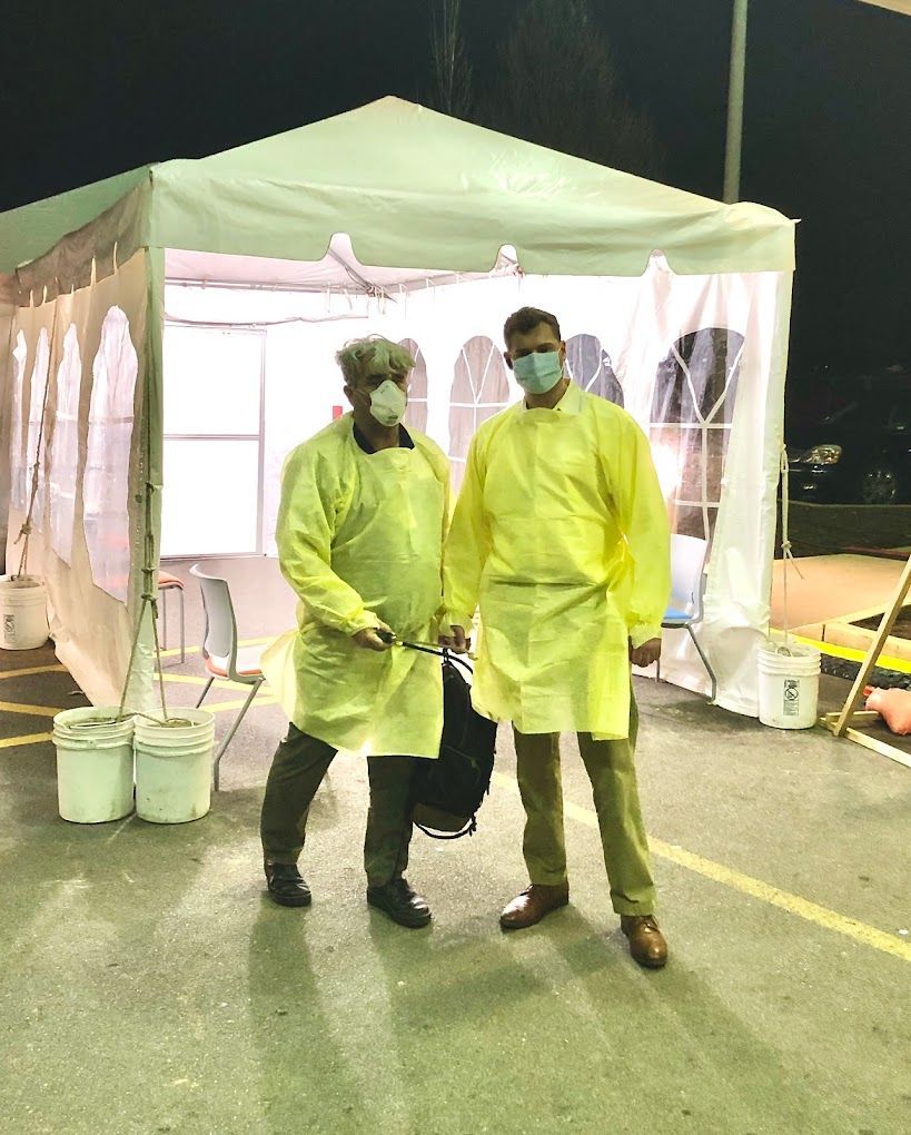 Two men wearing yellow gowns and masks are standing in front of a tent