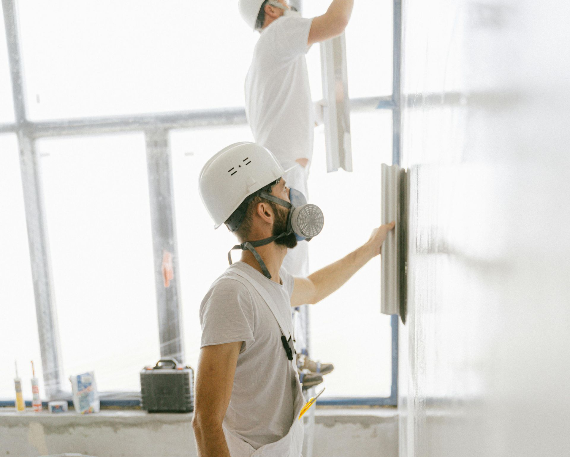 Two workers in white construction gear and helmets plaster a wall inside a building with large windows.