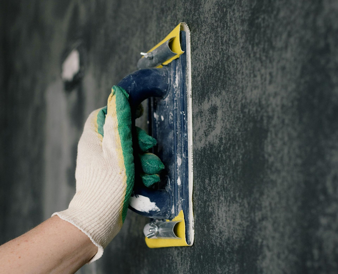 A gloved hand uses a blue and yellow sanding block to smooth a dark, textured wall surface.