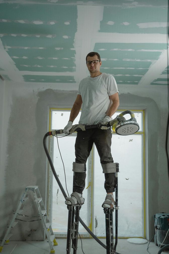 A person wearing stilts and protective gear stands in a room under construction, holding a ceiling sander.