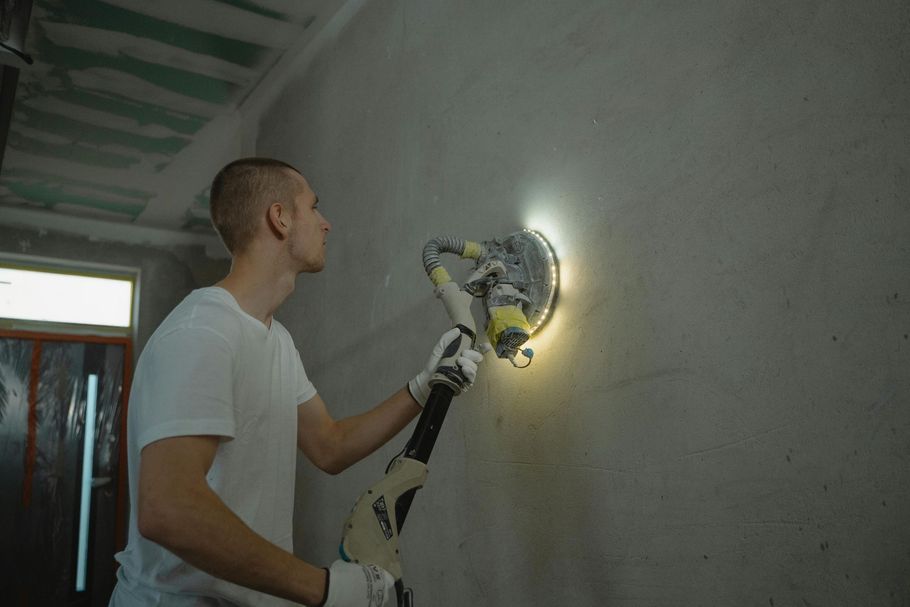 A person in a white t-shirt uses a handheld power sander with a light attachment to smooth a plaster wall.