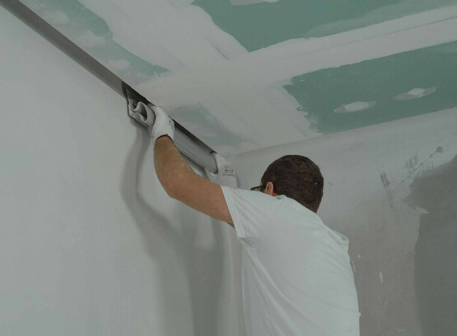 A person in a white shirt and gloves uses a tool to apply compound to the corner where a wall meets a drywall ceiling.