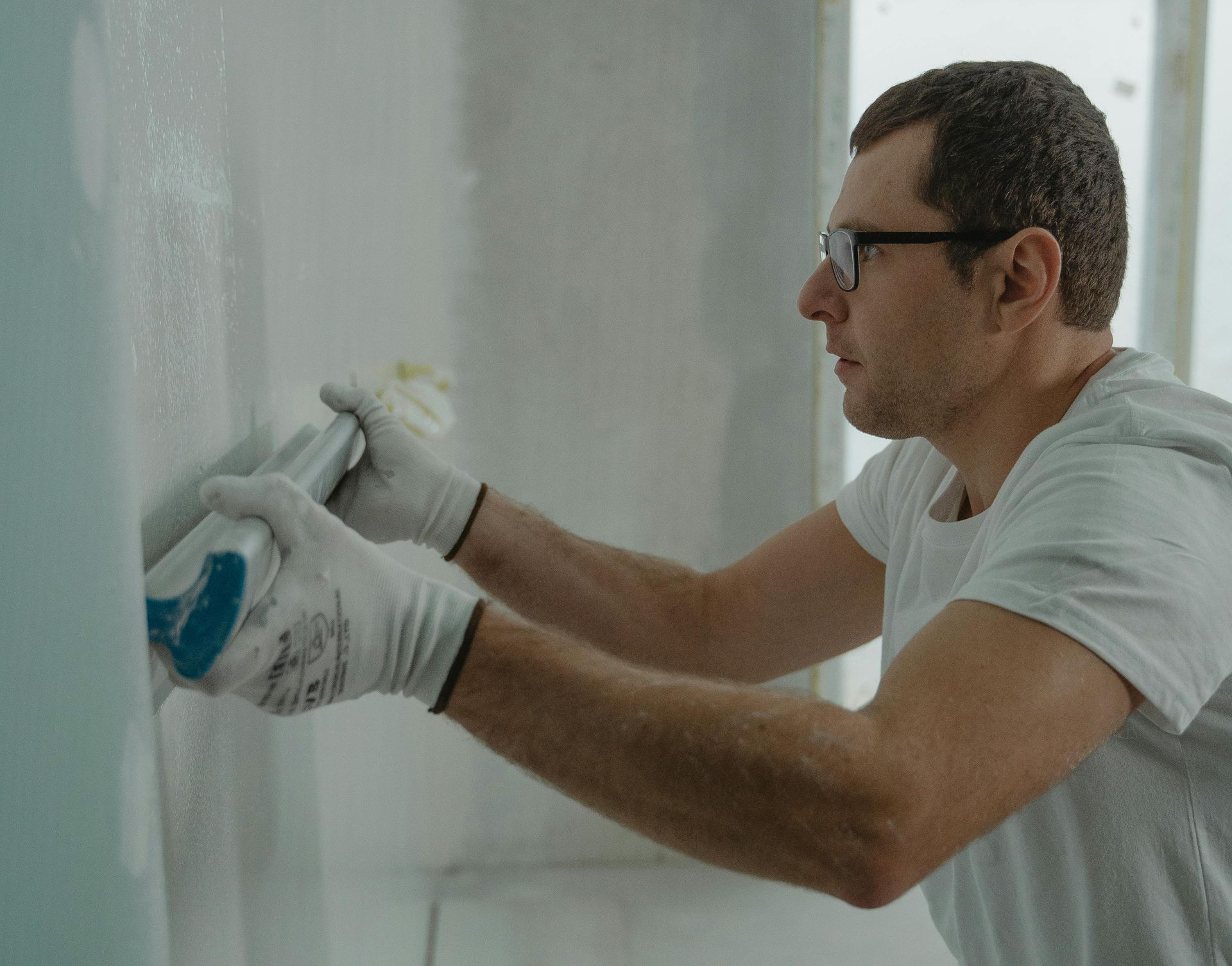 A person in white gloves and a white t-shirt uses a sanding block to smooth a white, plastered wall.