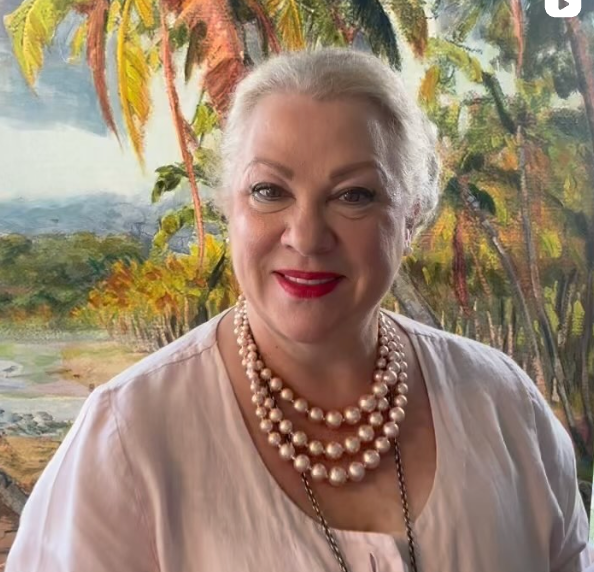 A woman wearing a pearl necklace is smiling in front of a painting of palm trees.