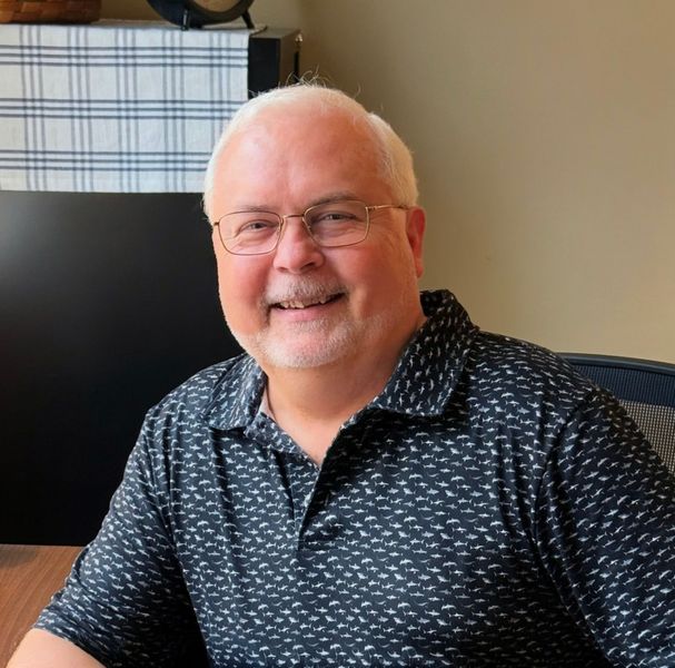 Smiling man with white hair and glasses, wearing a navy patterned shirt, seated indoors.