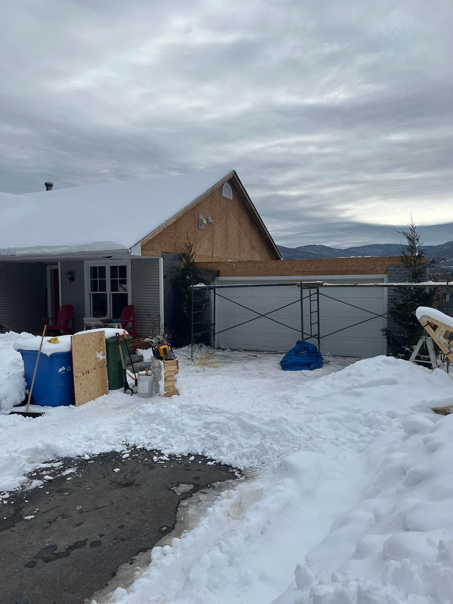 Chantier enneigé avec une maison en construction, divers matériaux et un ciel nuageux.
