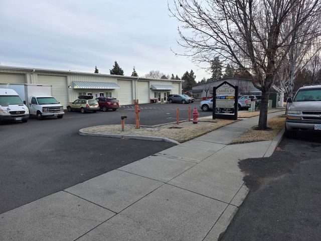 Sidewalk beside a parking lot with parked cars, a sign, bare trees, and a row of storefronts under cloudy sky | SP Auto and Transmission World