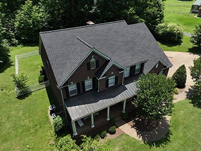Aerial view of a dark gray house with a porch, surrounded by green lawn and trees.