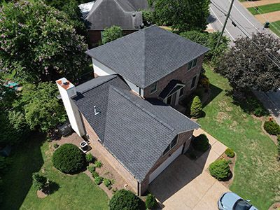 Aerial view of a gray-roofed brick house with a driveway and surrounding trees.