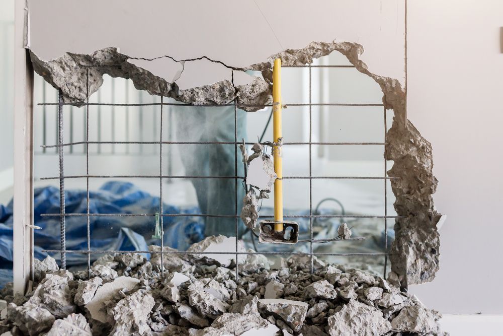 Damaged wall with a large hole revealing a room behind it and debris scattered on the floor.