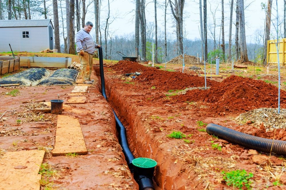 Trenched yard with black pipes and orange-red dirt at a construction site, worker in background