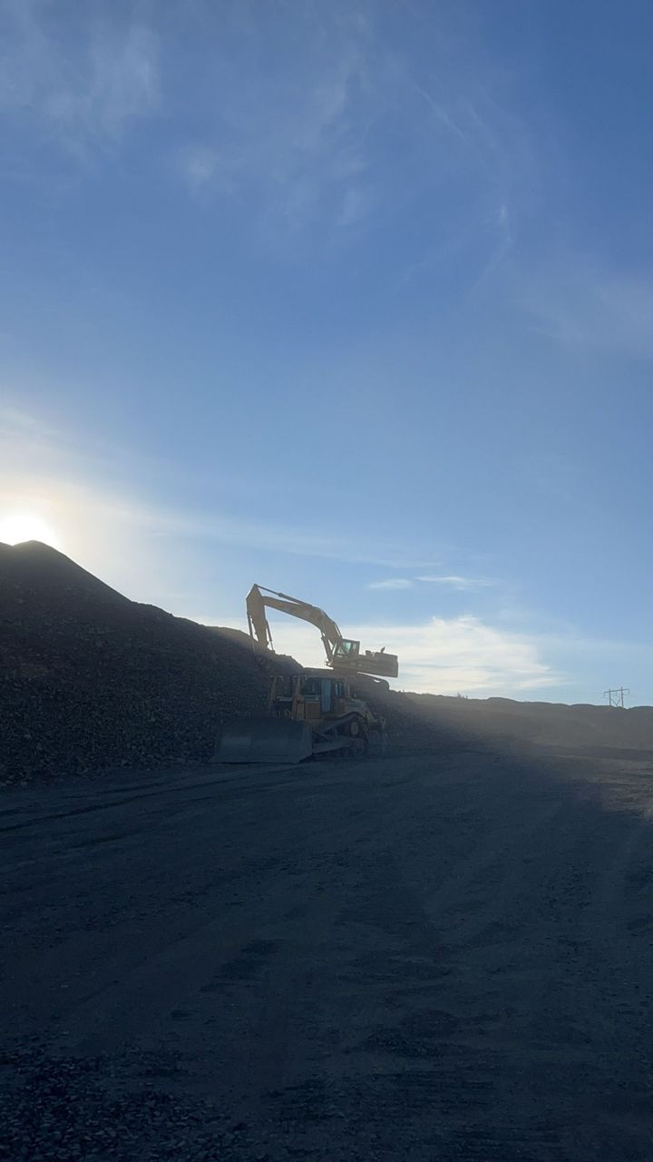 Excavator on a dark gravel slope at sunset, silhouetted against a blue sky.
