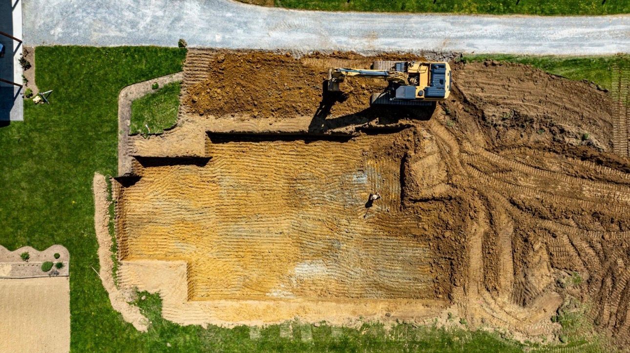 Aerial view of a bulldozer grading a dirt lot beside a green lawn and stone retaining wall.