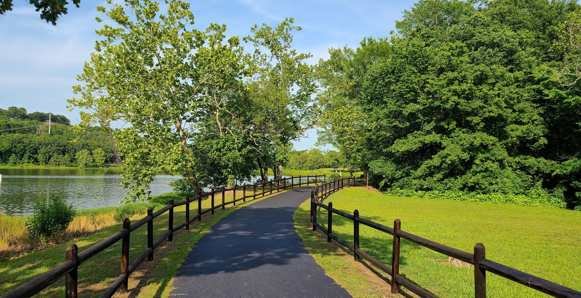 Paved path by a lake, lined with black fences and green trees on a sunny day