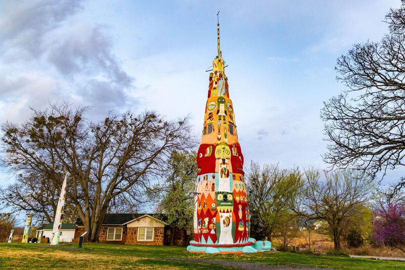 Tall colorful mosaic tower in a park with trees and houses under a cloudy sky