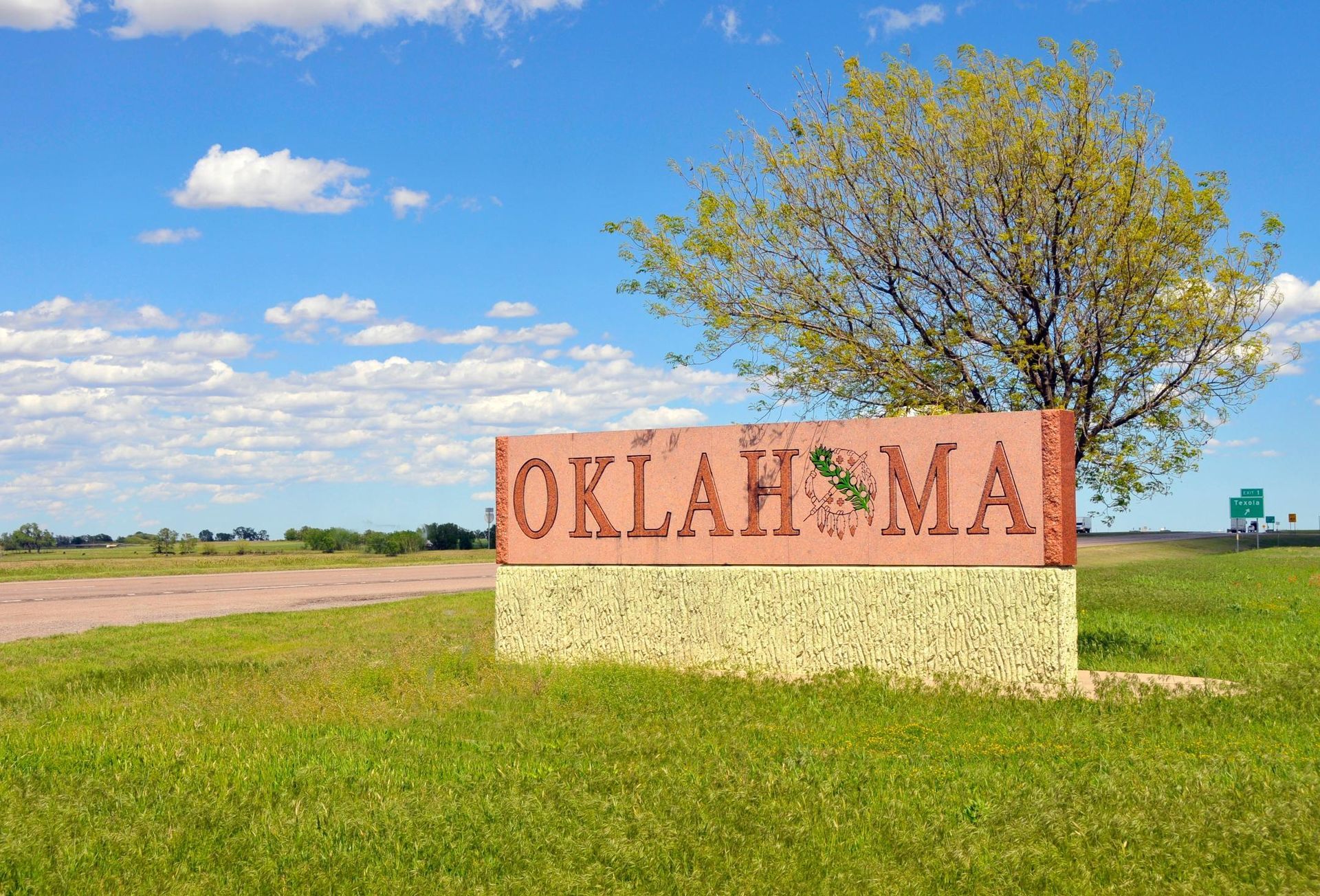 “OKLAHOMA” roadside sign on a grassy field under a blue sky with clouds