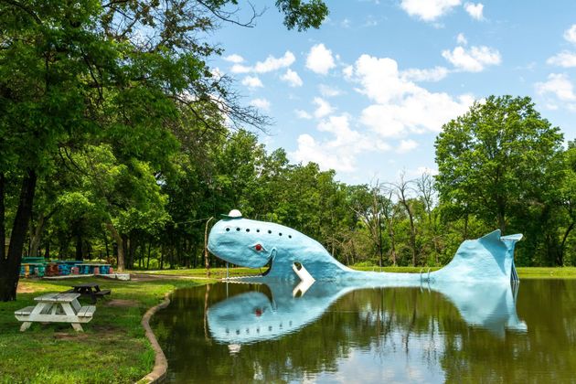 Large blue fish sculpture in a park pond, surrounded by trees and picnic tables under a sunny sky