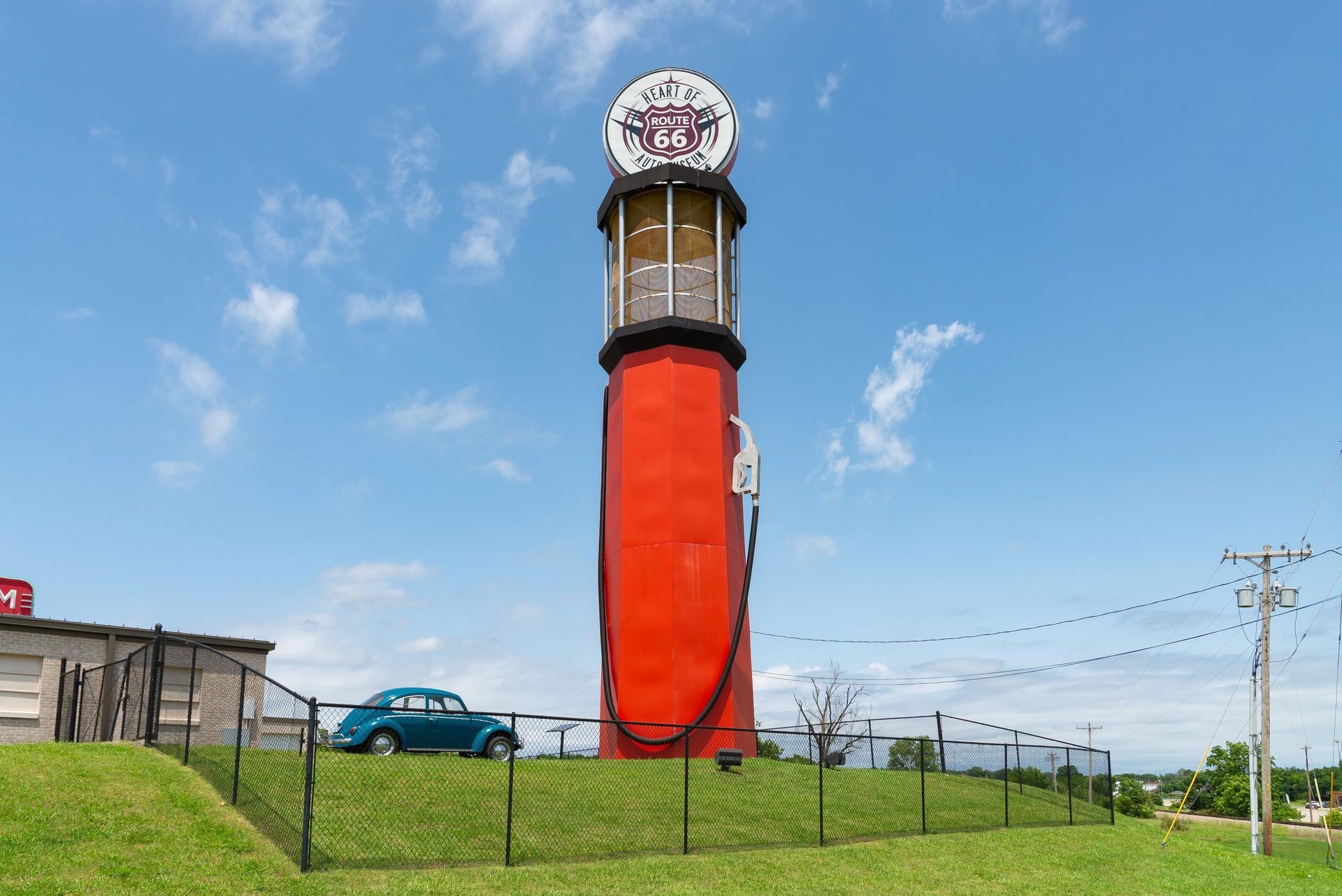 Tall red roadside clock tower with a large round sign on top, beside a grassy field and blue car under a clear sky