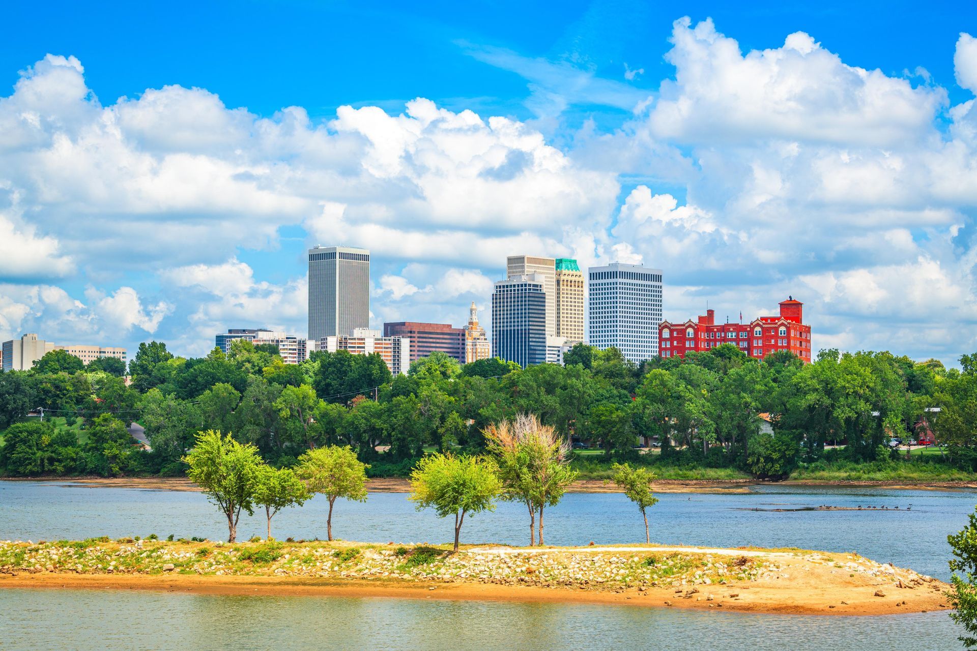 City skyline beyond a tree-lined river under a bright blue sky with white clouds