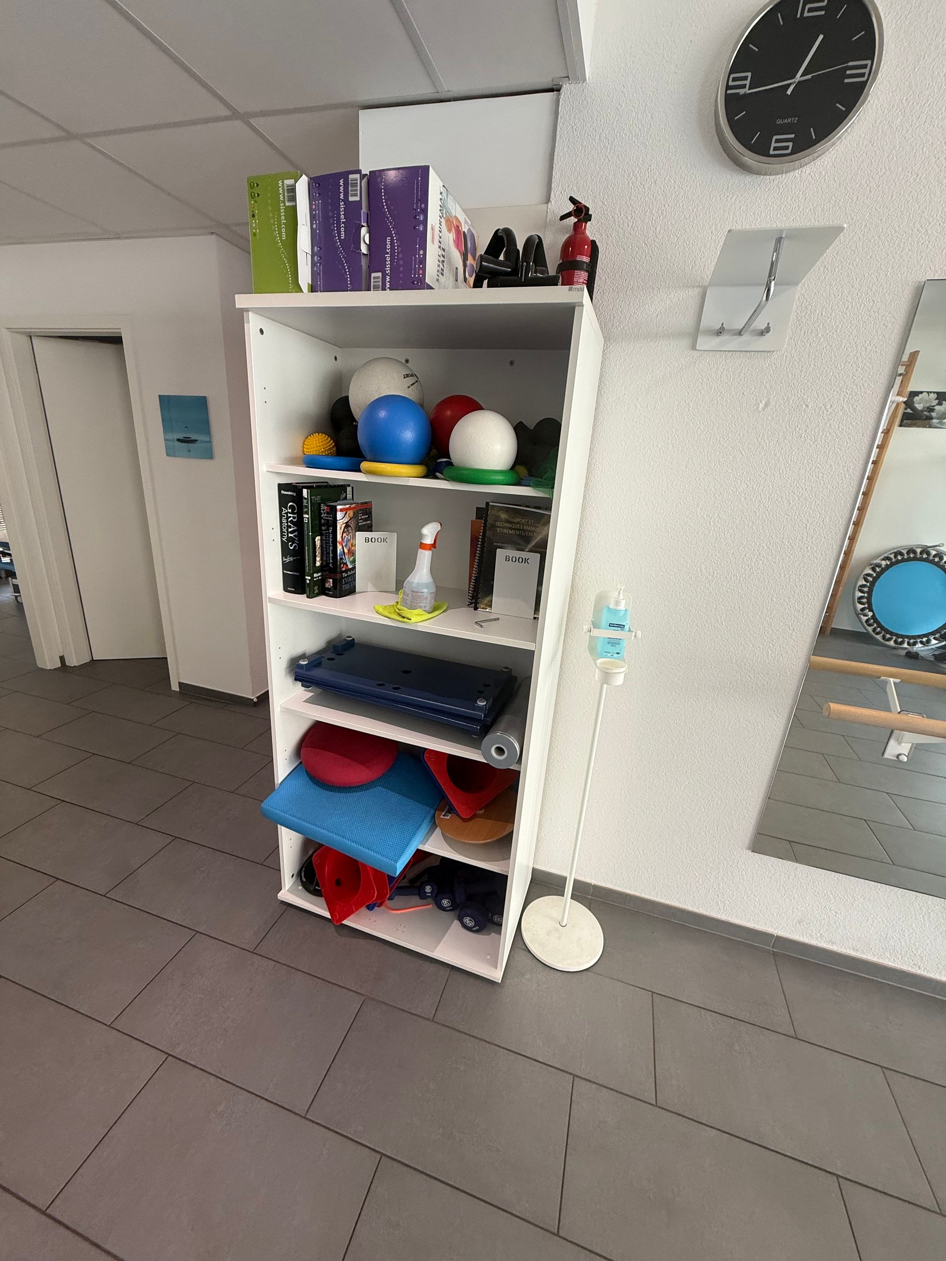 Étagère blanche avec des balles colorées et des fournitures, à côté d'un miroir et d'une horloge murale.