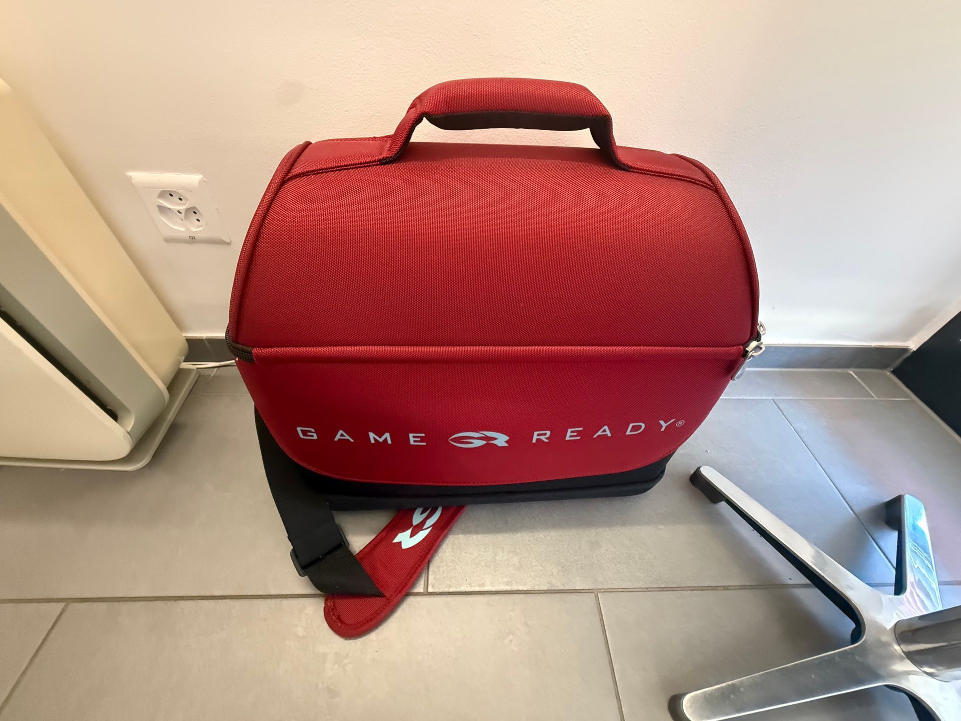 Sac rouge Game Ready à fermeture éclair avec poignée, posé au sol à côté d'une chaise et d'une prise murale.