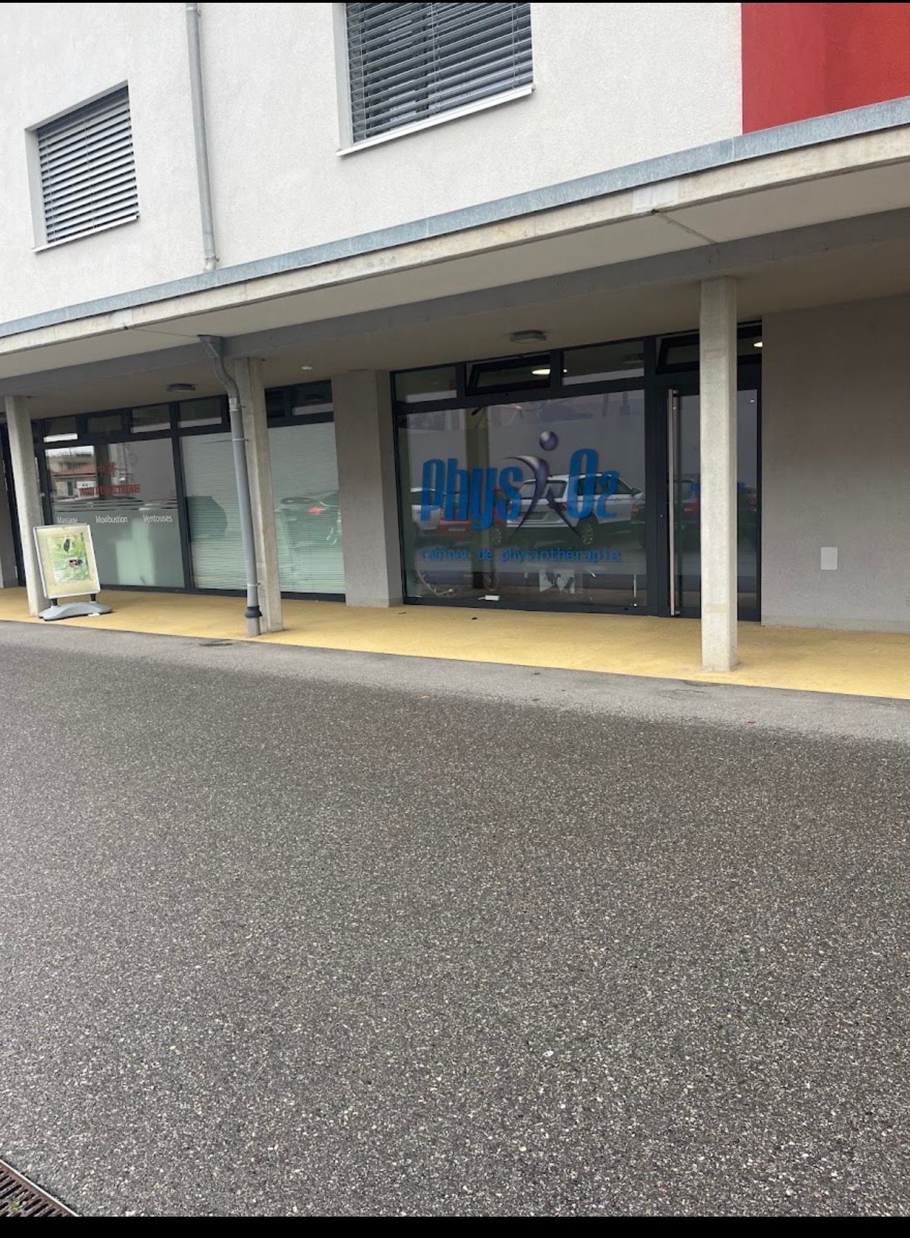 Entrée de magasin avec portes et fenêtres vitrées sous un trottoir couvert, visible depuis le parking.