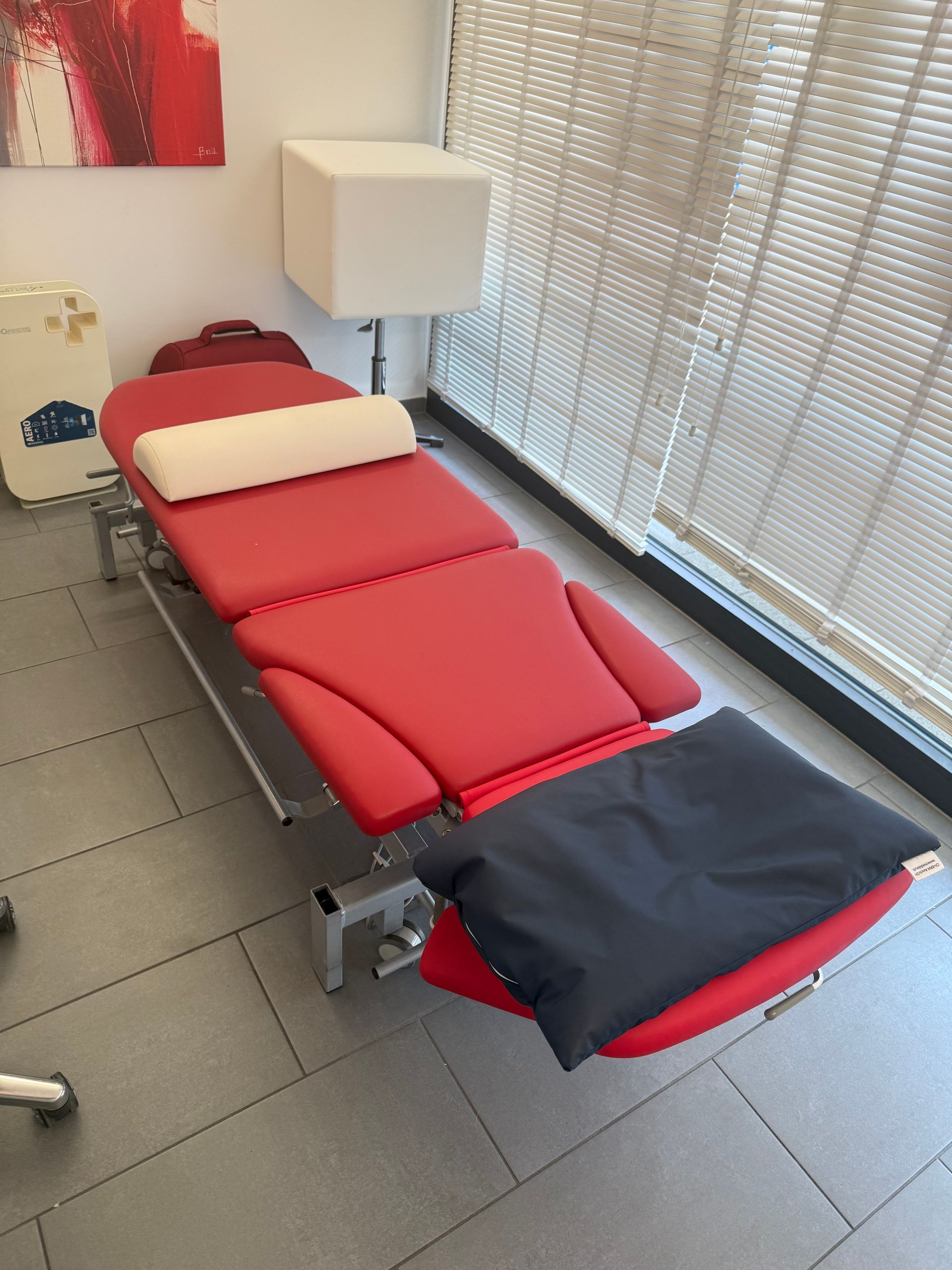 Table de soins rouge rembourrée dans une salle de clinique, à côté d'une fenêtre à stores.