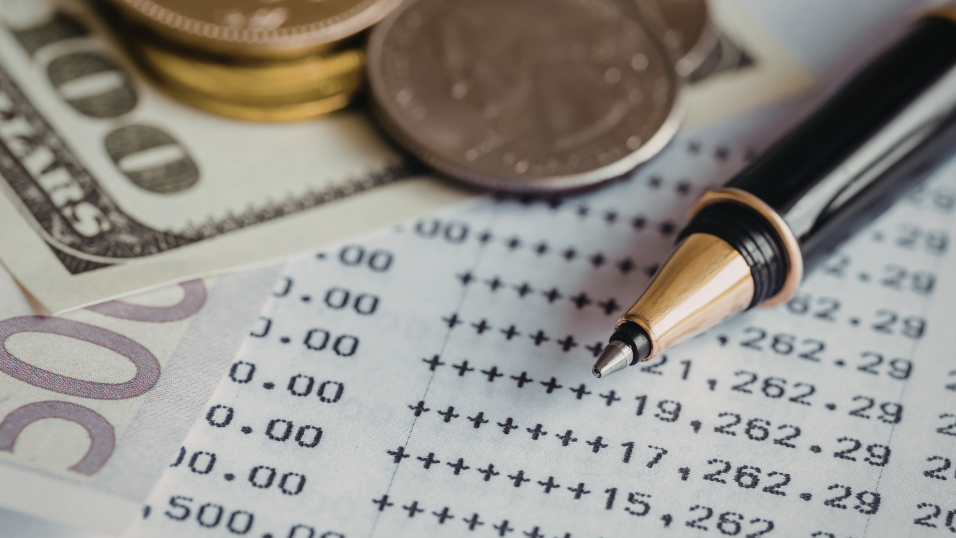 A pen rests on a financial ledger with rows of numbers, with US currency and coins visible in the background.