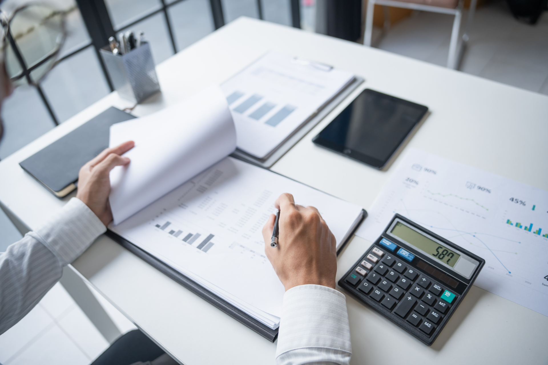 A person in a light-colored shirt analyzing financial documents and bar charts at a desk with a calculator and tablet.