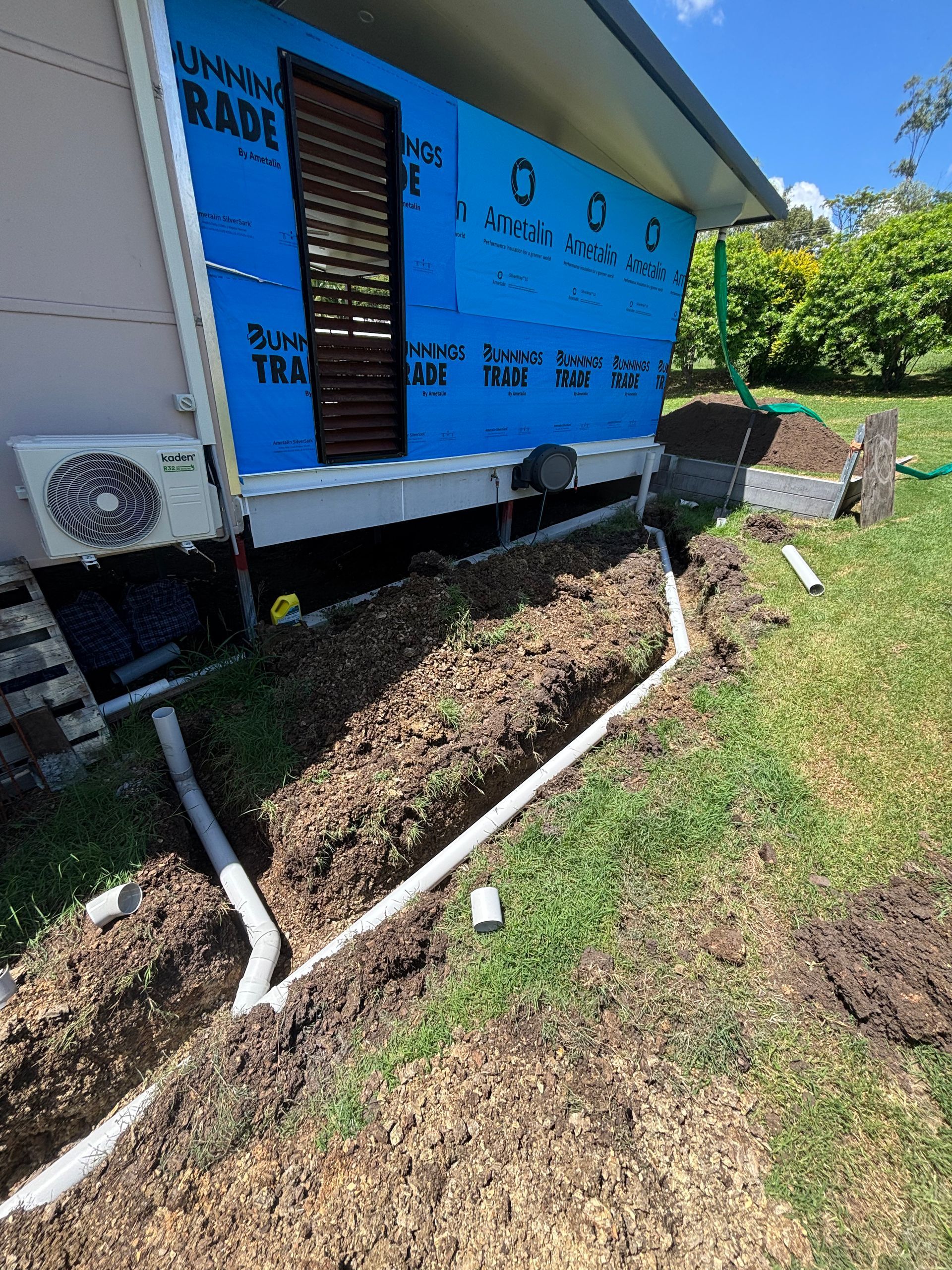 Blue house exterior with exposed pipes, an AC unit, and a dirt trench along the foundation — Truflow Plumbing & Gas Solutions in Rockhampton City, QLD