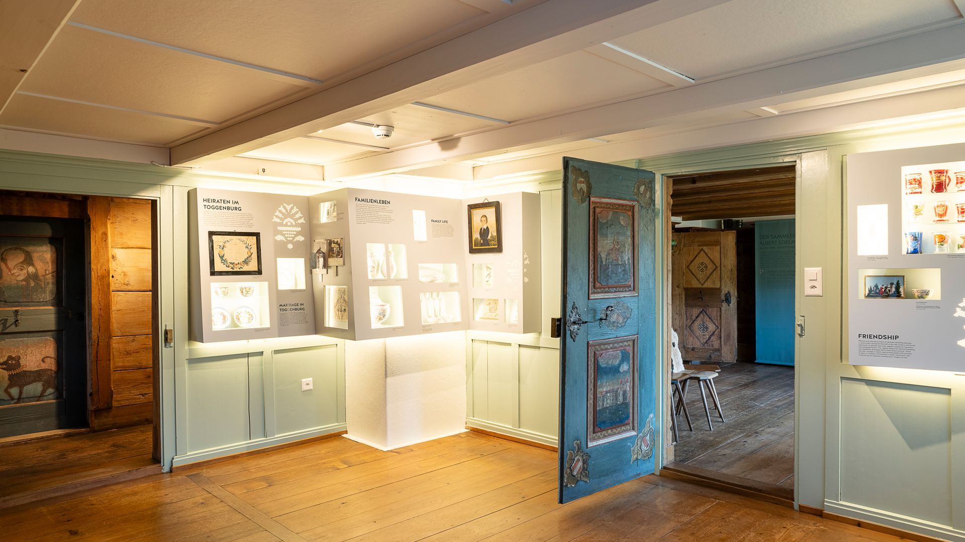 Museumsraum im Ackerhus mit Ausstellungsstücken, Holzfußboden, blau-weißen Zierleisten, offene Tür zu einem anderen Raum.
