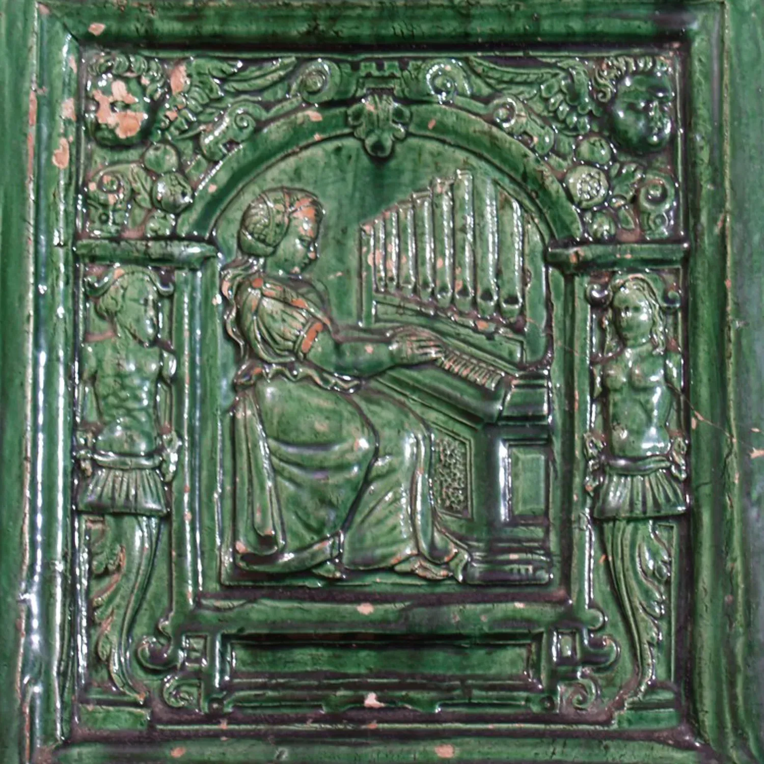 Relief einer Frau, die Orgel spielt, auf grünen Keramikfliesen, flankiert von Figuren und floralen Motiven.