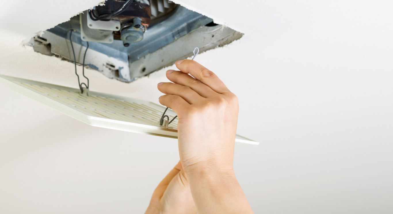 Person's hands attaching a white, square cover to a ceiling ventilation unit. Wires are visible.
