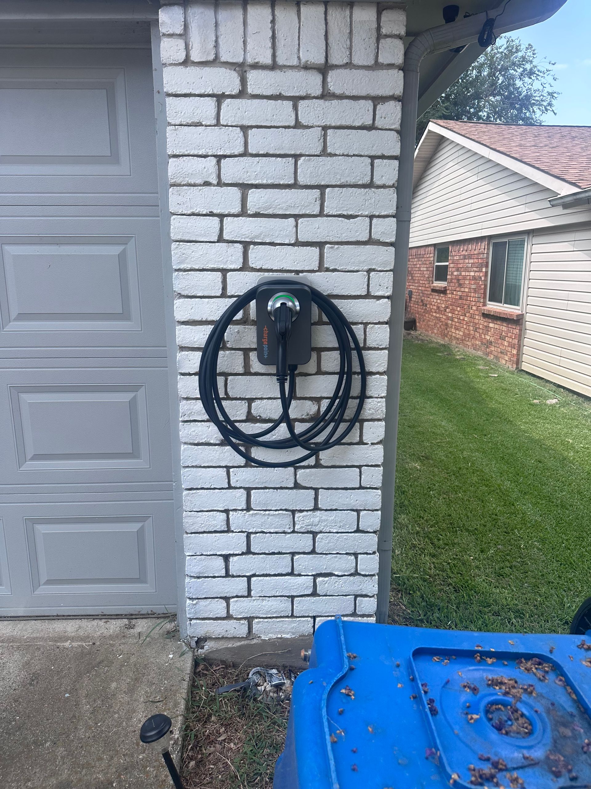 Electric vehicle charger mounted on a brick wall next to a garage door. Blue recycling bin visible below.