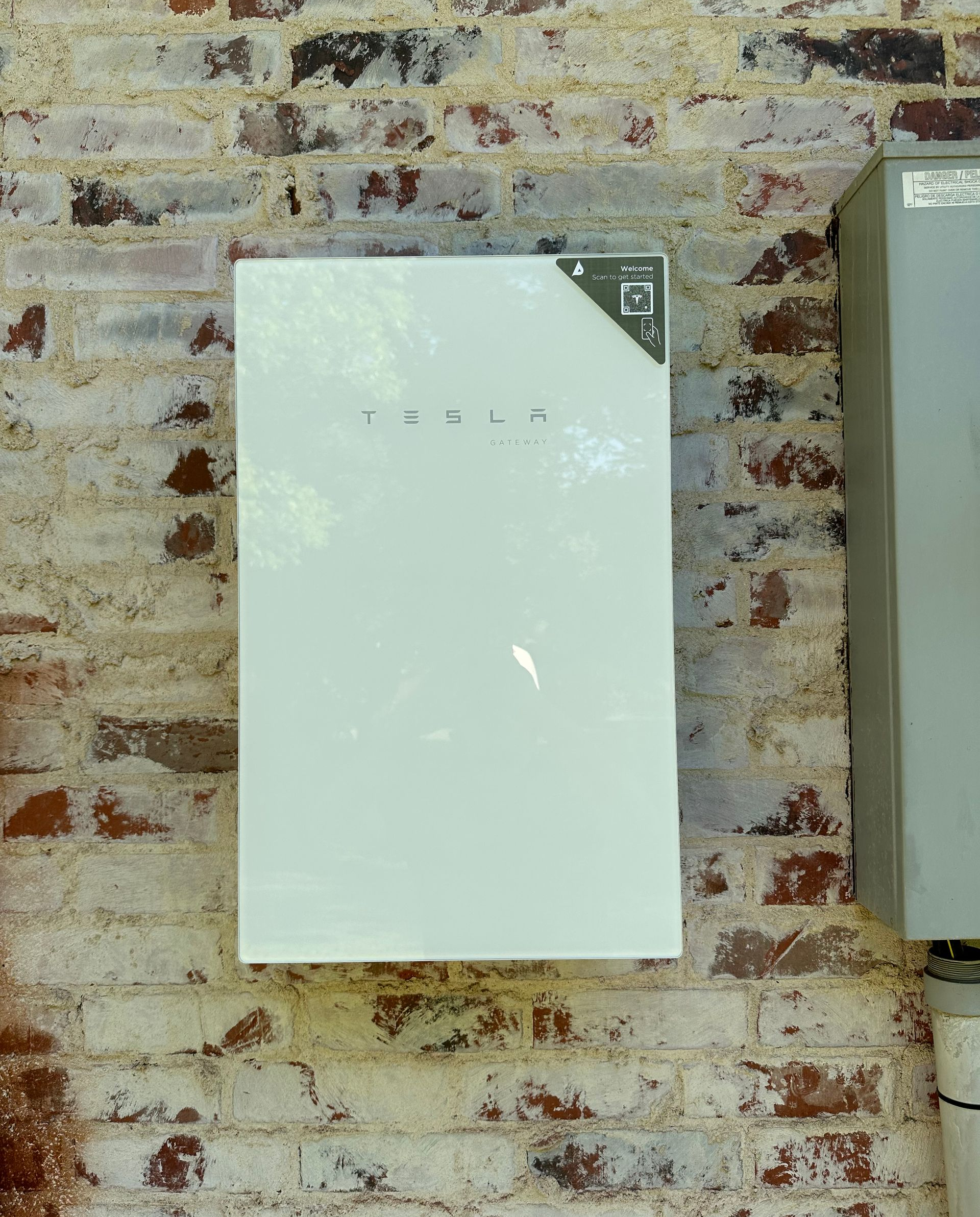 White Tesla Powerwall mounted on a weathered brick wall next to a gray electrical box.