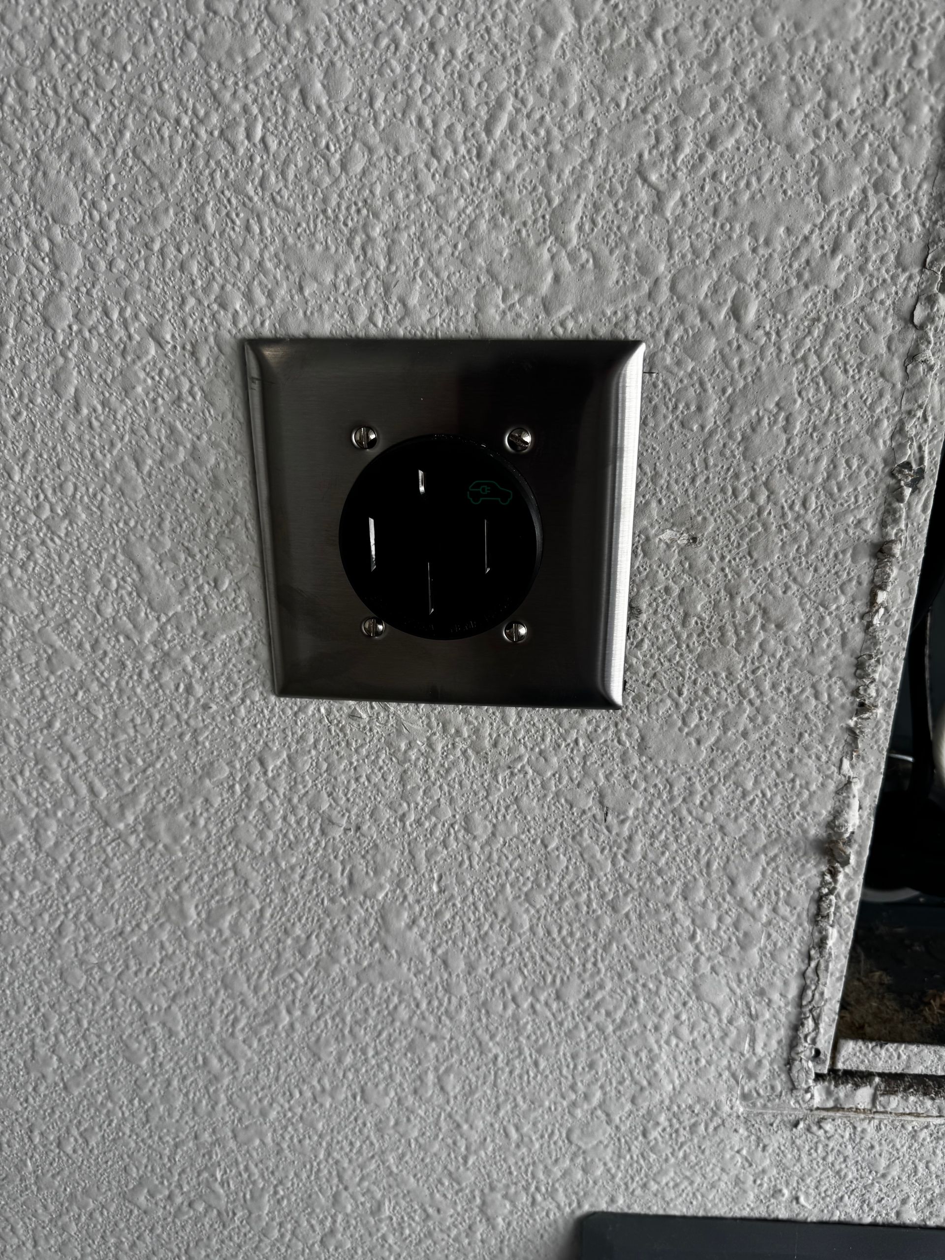 Black, round 240-volt electrical outlet with three prongs set in a square silver metal plate on a textured, gray wall.