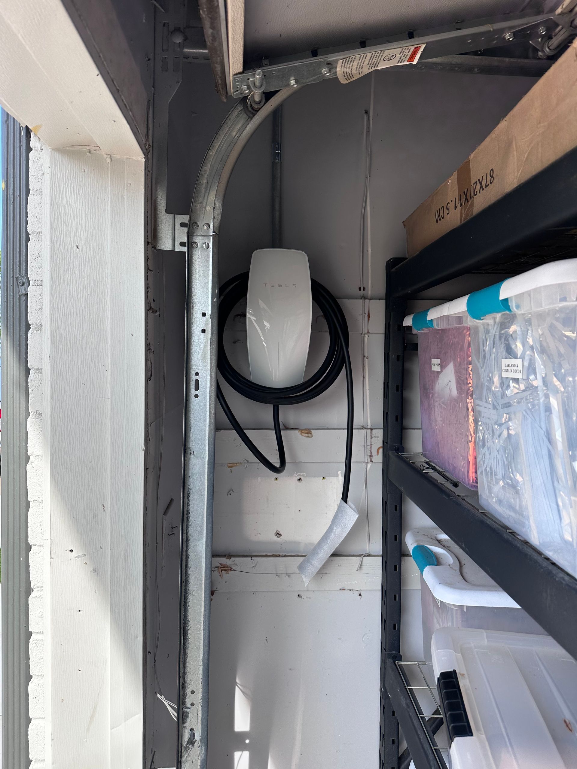 Electric vehicle charger mounted in a garage near shelves holding storage bins.